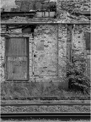 Verfallenes Mauerwerk mit einer Holztür, umgeben von Gras und Gleisen, in Schwarz-Weiß.