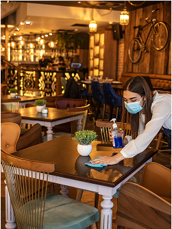 Eine Restaurantmitarbeiterin mit Maske reinigt einen Tisch in einem stilvollen Restaurant.