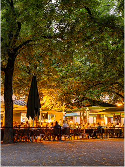 Gemütliches Freiluftrestaurant unter Bäumen mit beleuchteten Tischen und Stühlen in warmem Licht.