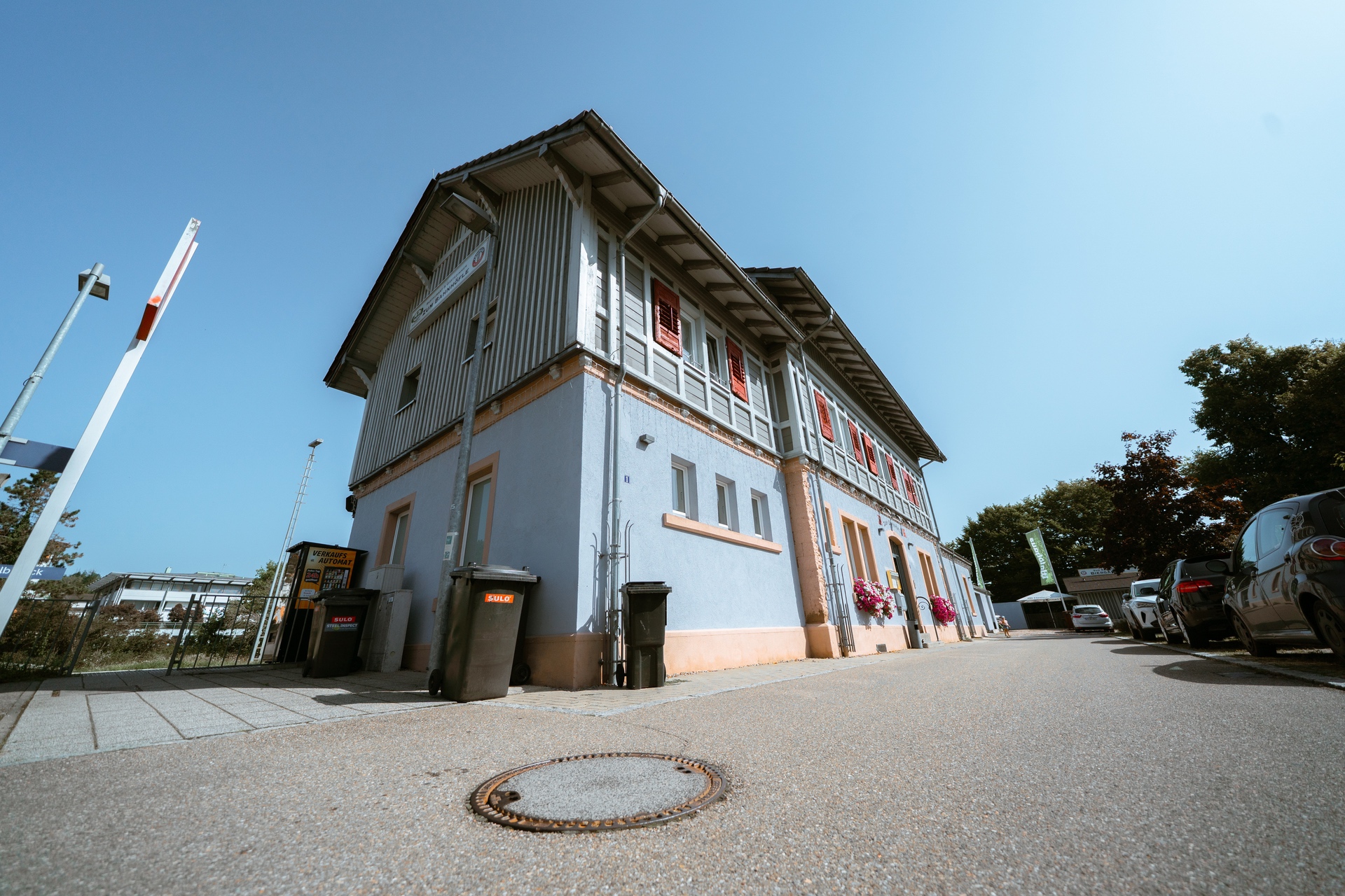 Ein hellblaues Gebäude mit roten Fenstern, Mülltonnen, und einem klaren Himmel darüber. Parkplatz auf der Seite.
