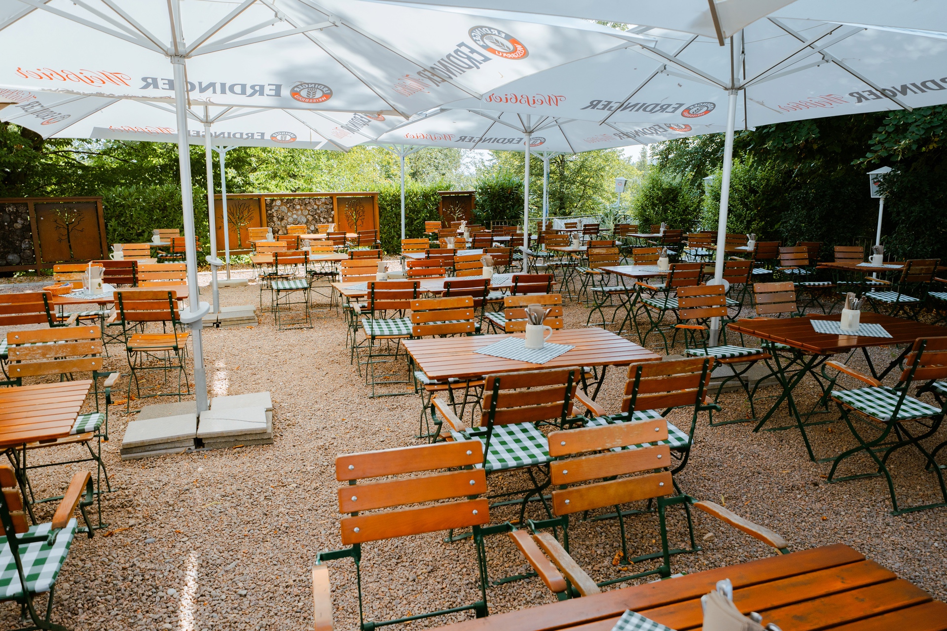Ein leerer Gartenbereich mit Holzstühlen und Tischen, überdacht von großen Sonnenschirmen.