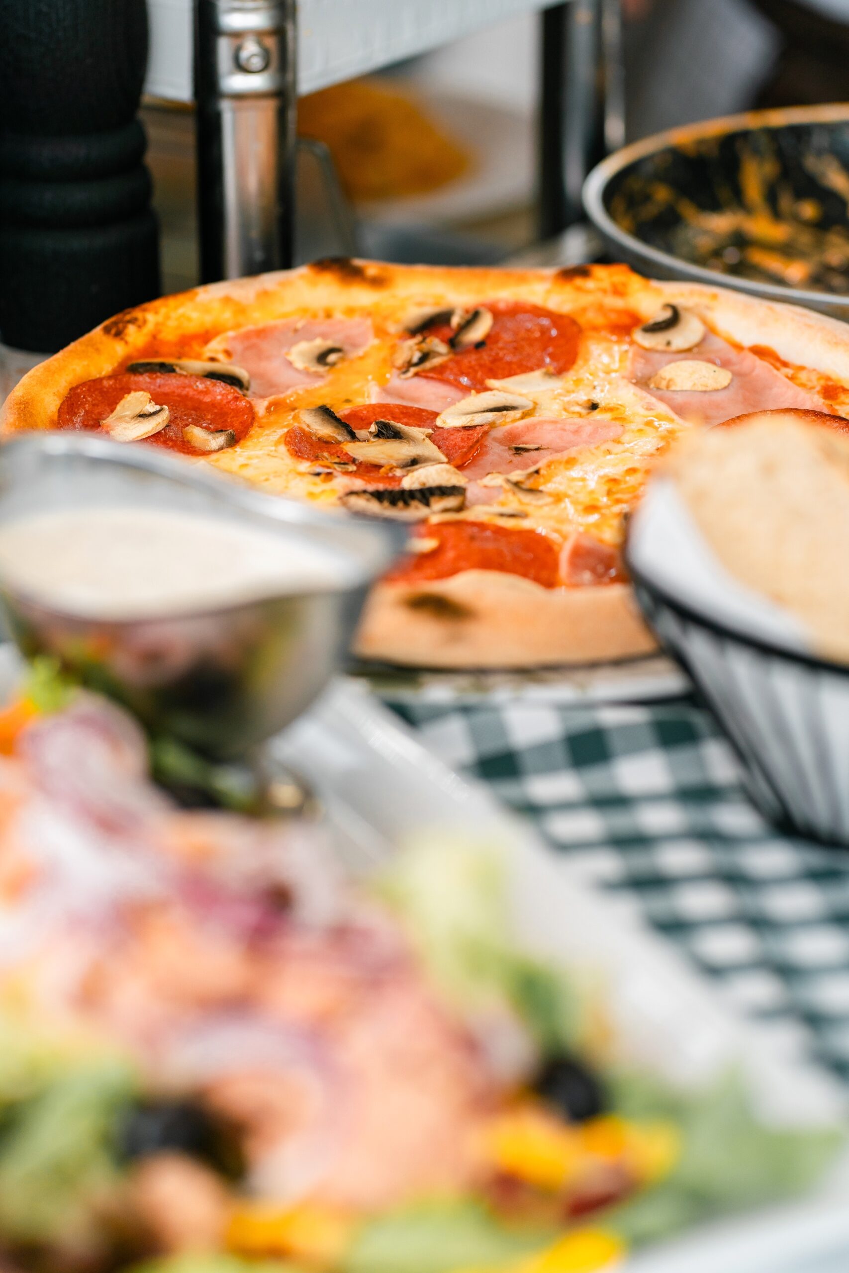 Eine Pizza mit Salami, Schinken und Pilzen, umgeben von Salat und Saucen in einem Restaurant.