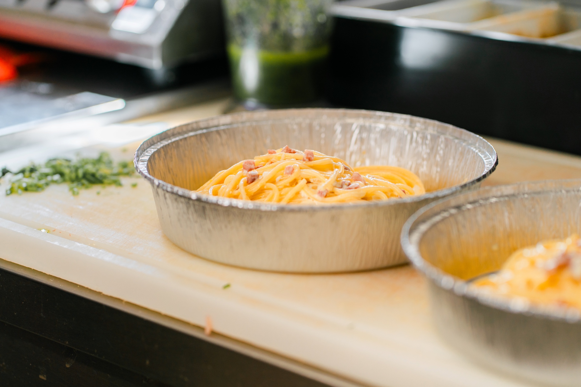 Ein Aluschalen mit Spaghetti in einer Küche, neben frischen Kräutern auf einem Schneidebrett.
