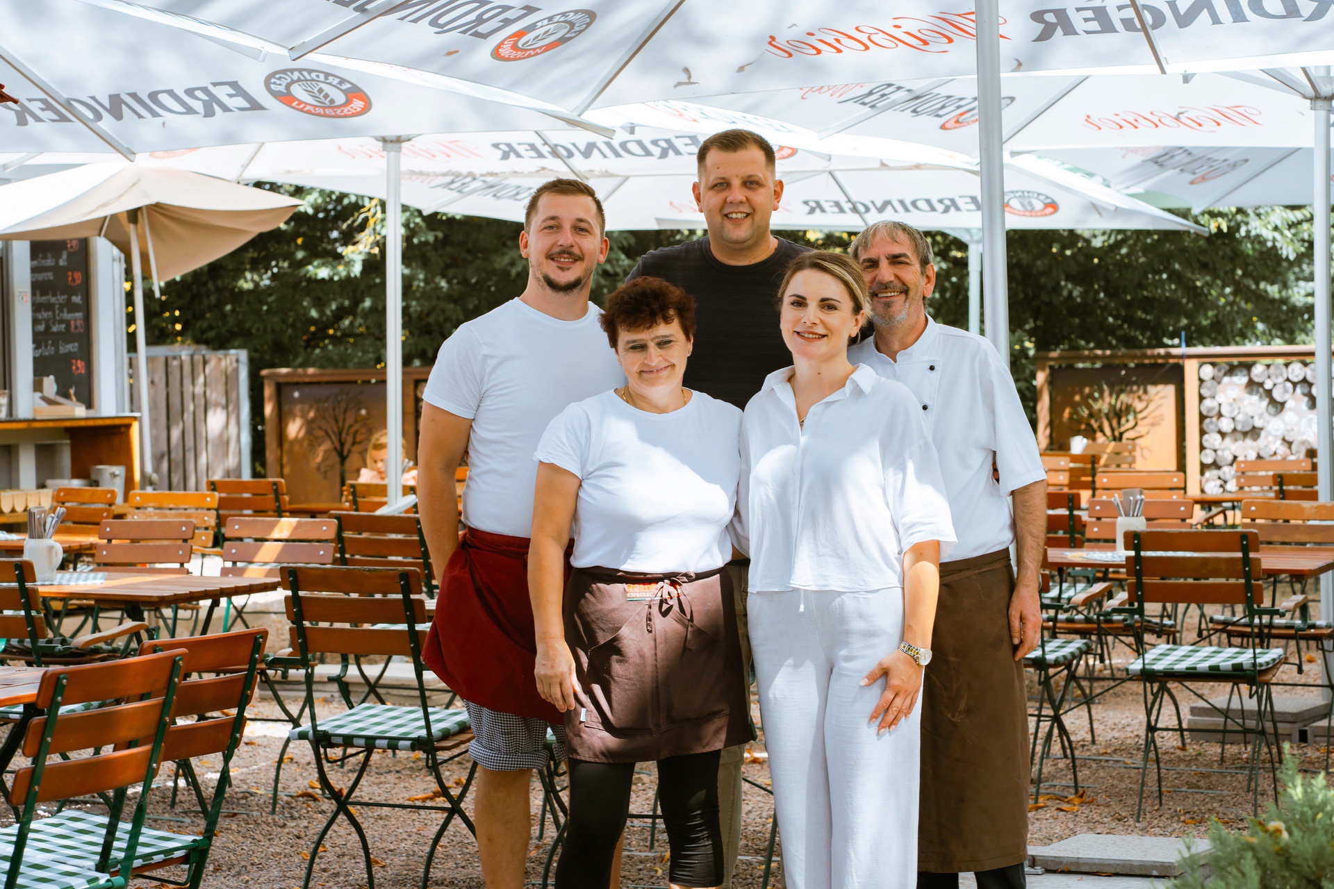 Fünf Mitarbeitende stehen lächelnd in einem Restaurantgarten unter Sonnenschirmen. Tische und Stühle sind sichtbar.